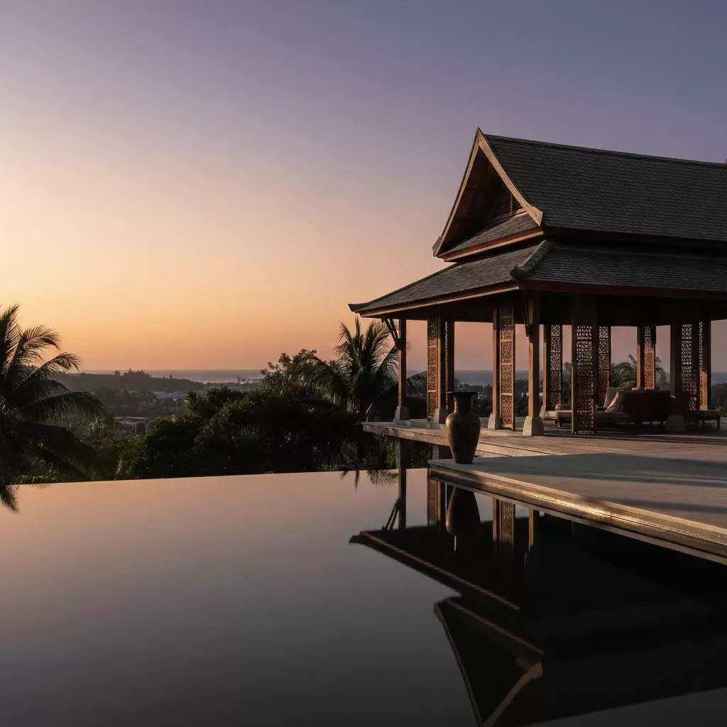 Thai-style pavilion with carved teak screens beside a still infinity pool at sunset, palms and sea beyond.