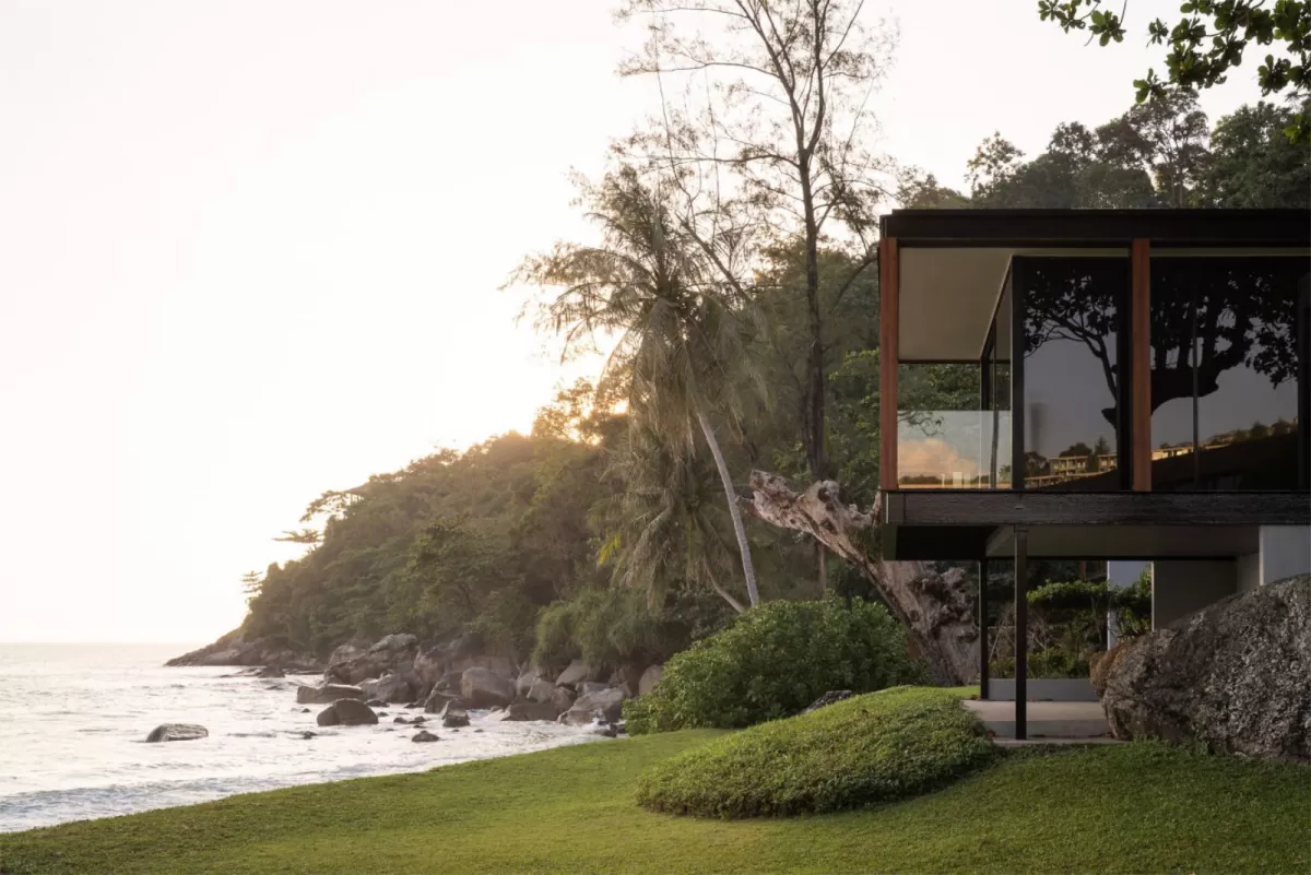 Glass-walled villa raised above a lawn on a rocky shoreline at sunset, with trees and sea alongside.