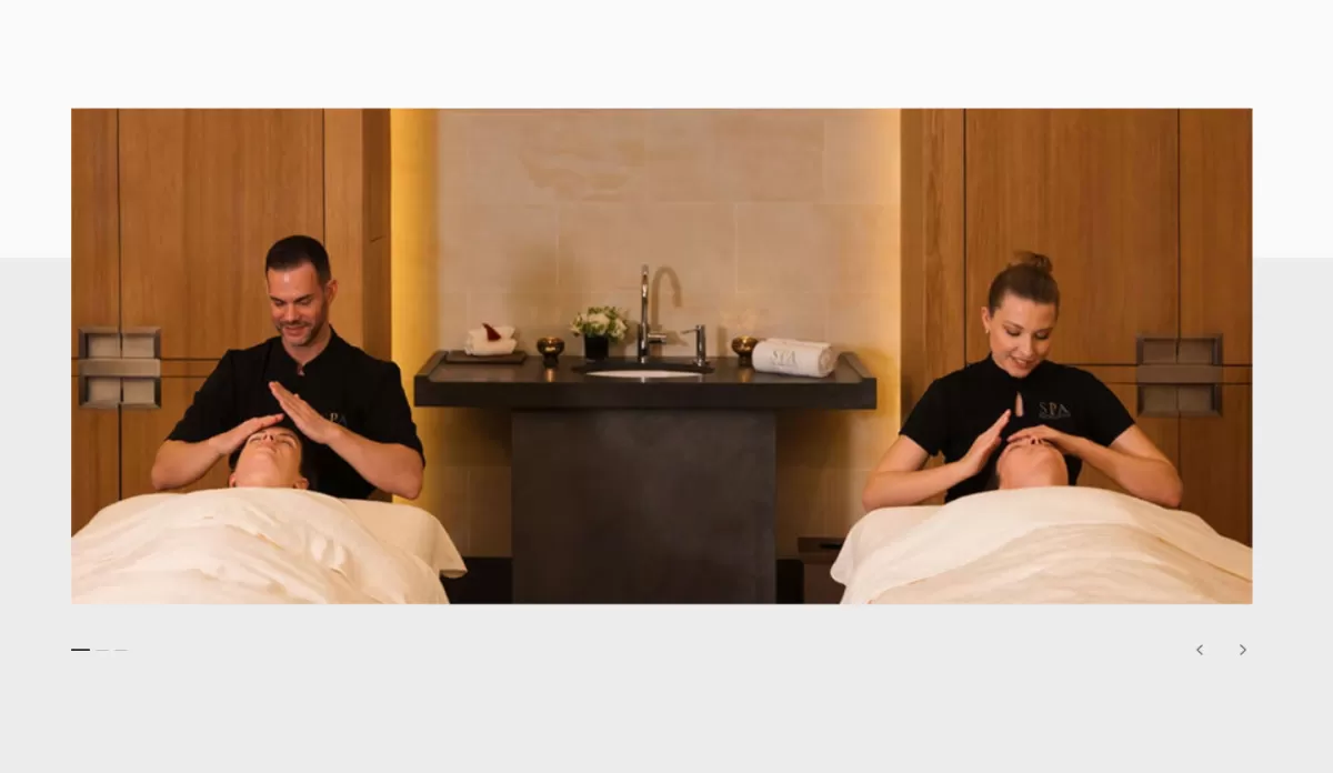 Two professional spa therapists concurrently administering facial massages to two guests lying down on parallel massage beds inside a luxury wellness suite.