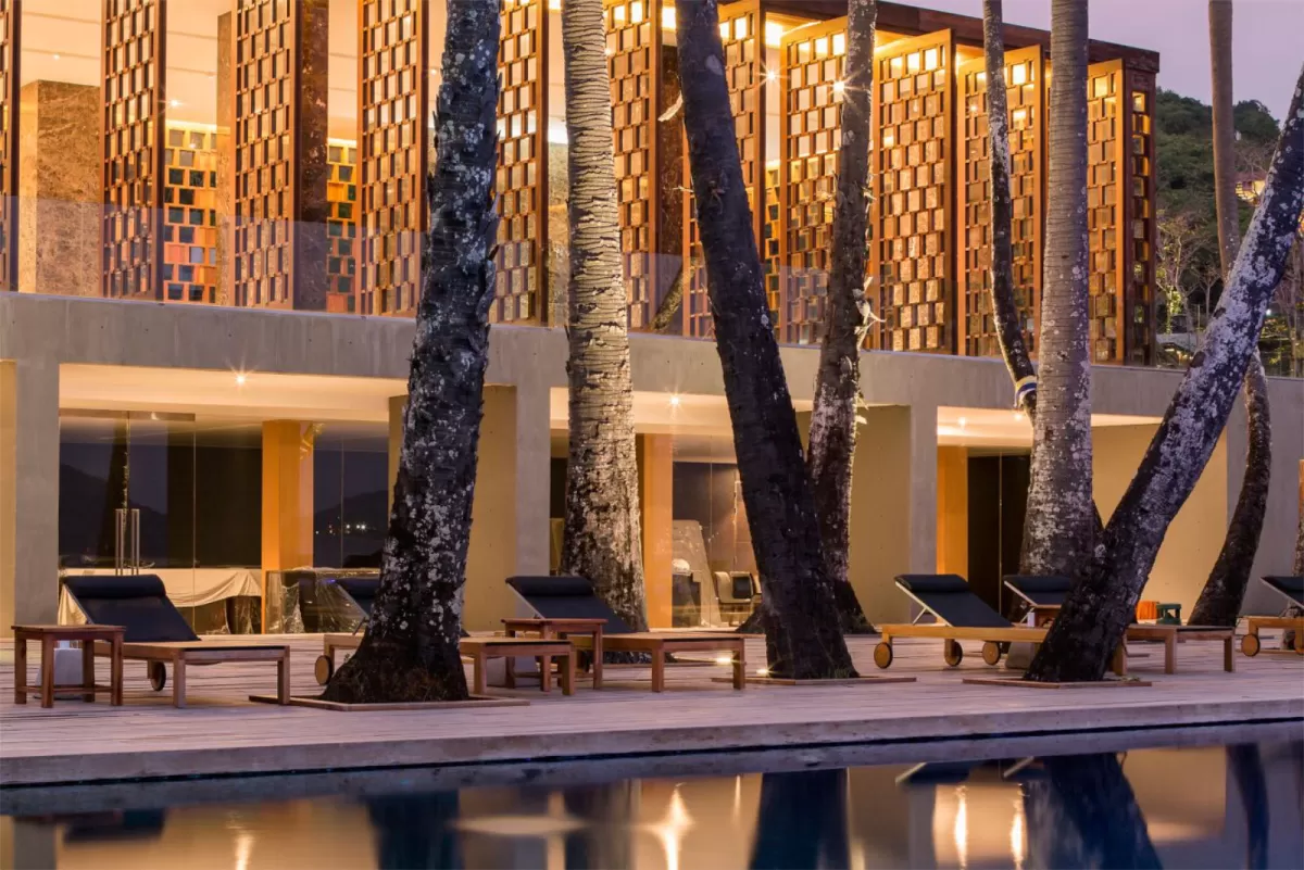 An evening view of a luxury resort pool deck lined with wooden lounge chairs positioned beneath rows of tall, lit palm trees in front of a modern architectural facade with geometric wood-screened paneling.