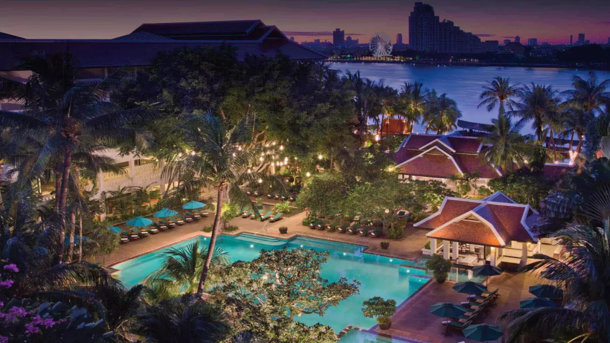 Elevated evening view of a luxury riverside resort featuring a large lit swimming pool surrounded by tropical palm trees, lounge chairs with umbrellas, and traditional pavilions along a wide river with a city skyline and illuminated Ferris wheel in the background.